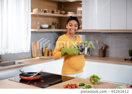 Smiling young pregnant african american woman with big belly in protective gloves prepares dinner 93261612