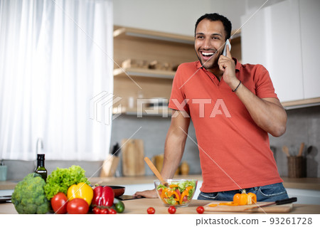 Smiling millennial african american guy preparing salad with organic vegetables speaks by smartphone 93261728