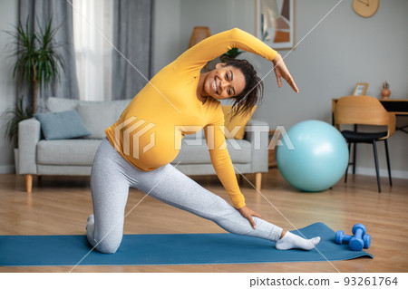 Happy smiling young pregnant black female practicing yoga, stretching body on mat in living room interior 93261764
