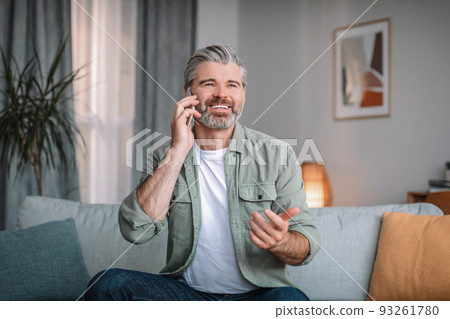 Cheerful caucasian senior male with beard calls by phone in living room interior. Retired talking to client at home Cheerful caucasian senior male with beard calls by phone in living room interior. Retired talking to client at home 93261780