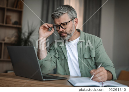 Busy serious european mature man teacher with beard in glasses looks at laptop, makes notes in room Busy serious european mature man teacher with beard in glasses looks at laptop, makes notes in room 93261840
