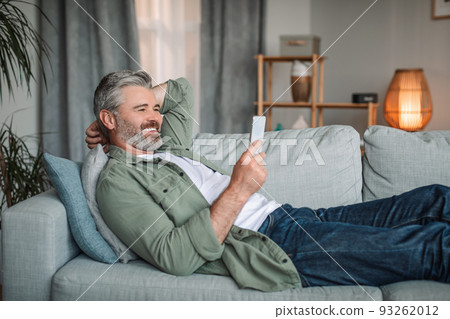 Cheerful caucasian old man with beard watch video on smartphone, rest and relax at sofa alone in living room Cheerful caucasian old man with beard watch video on smartphone, rest and relax at sofa alone in living room 93262012
