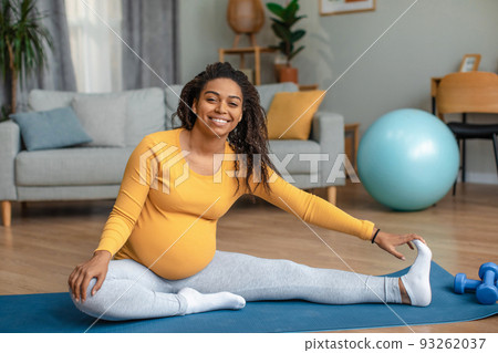 Happy young pregnant african american woman with big belly doing exercises and practicing yoga in living room 93262037