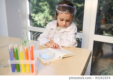 View from above of a smart school child, little Caucasian girl in protective lab wear reading chemistry textbook, sitting at desk with labware and rainbow color chemicals in test tubes on tripod View from above of a smart school child, little Caucasian girl in protective lab wear reading chemistry textbook, sitting at desk with labware and rainbow color chemicals in test tubes on tripod 93262834