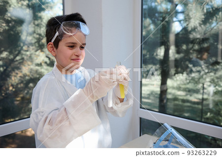 Caucasian schoolboy in lab coat and safety goggles, learns about science, holds test tubes, watches fascination going on chemical reaction, standing against panoramic windows in school laboratory Caucasian schoolboy in lab coat and safety goggles, learns about science, holds test tubes, watches fascination going on chemical reaction, standing against panoramic windows in school laboratory 93263229
