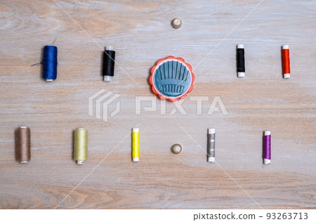 A plastic box with a set of sewing needles and many spools of thread of different colors are laid out in rows on a wooden textured surface as examples of needlework items. A plastic box with a set of sewing needles and many spools of thread of different colors are laid out in rows on a wooden textured surface as examples of needlework items. 93263713