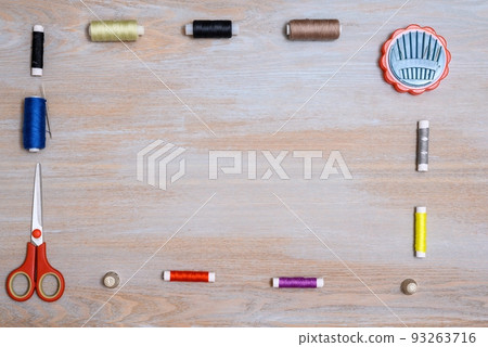 Scissors as well as many spools of thread with needles and needlework kits are laid out along the edge of a horizontal wooden surface forming a frame for tex. 93263716