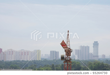 One construction crane while working at a construction site in cloudy spring weather against the backdrop of a blue sky, with clouds and multi-storey buildings and far forest as a background. One construction crane while working at a construction site in cloudy spring weather against the backdrop of a blue sky, with clouds and multi-storey buildings and far forest as a background. 93264730