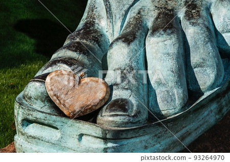 A hand-crafted wooden heart with a rustic rough structural and textured surface rests beside a patina-covered foot of a Greek sandal-clad bronze monument situated in a lawn of green grass. 93264970