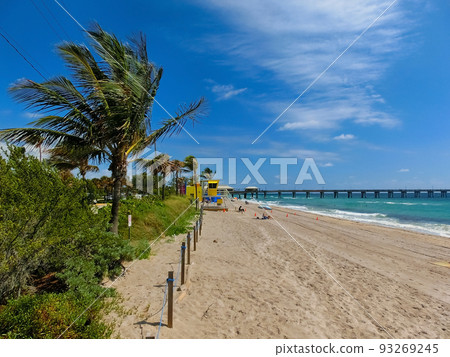 Long distance view of the sunny and tropical blue water shoreline with people, foliage, boats and buildings in the background seen from above the water. 93269245