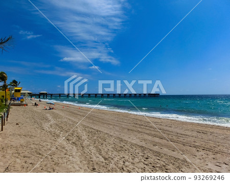Long distance view of the sunny and tropical blue water shoreline with people, foliage, boats and buildings in the background seen from above the water. Long distance view of the sunny and tropical blue water shoreline with people, foliage, boats and buildings in the background seen from above the water. 93269246