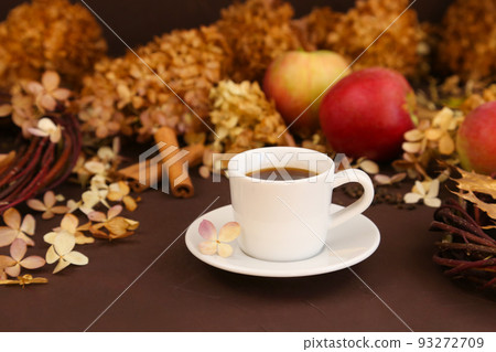 White coffee cup, autumn leaves, cinnamon, apples and dry hydrangea on brown background, front view. Selective focus White coffee cup, autumn leaves, cinnamon, apples and dry hydrangea on brown background, front view. Selective focus 93272709