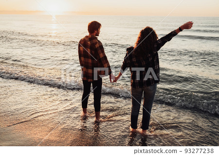 Young couple meeting sunrise on the seashore, man and woman holding by hands standing in water Young couple meeting sunrise on the seashore, man and woman holding by hands standing in water 93277238