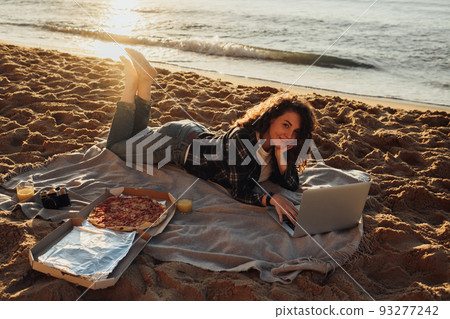 Happy curly haired woman eating pizza and working on laptop while laying on the seashore at dawn Happy curly haired woman eating pizza and working on laptop while laying on the seashore at dawn 93277242