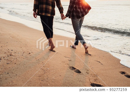 Unrecognisable couple meeting sunrise on the seashore, man and woman holding by hands walking along the coastline Unrecognisable couple meeting sunrise on the seashore, man and woman holding by hands walking along the coastline 93277259