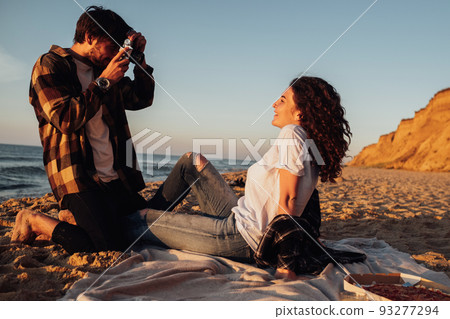 Young man photography his curly haired girlfriend that sitting on the seashore at dawn, couple enjoying sunrise on the beach Young man photography his curly haired girlfriend that sitting on the seashore at dawn, couple enjoying sunrise on the beach 93277294