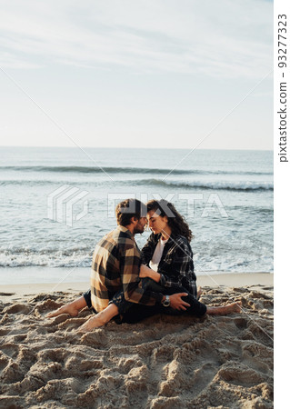 Young woman and man hugging while sitting together on the seashore with sea waves on the background at sunrise Young woman and man hugging while sitting together on the seashore with sea waves on the background at sunrise 93277323