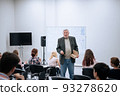A male teacher in a jacket is giving a lecture on mathematics at the university, he is standing at the white board, students are sitting in a large audience on chairs and listening attentively 93278620