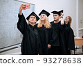 Young happy graduates of the academy take selfies on a smartphone in academic gowns and caps and smile 93278638