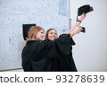 During the presentation of graduation diplomas at the university, two beautiful girls in academic gowns and caps take selfies and smile 93278639