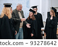 In the math class, an elderly gray-haired professor congratulates students in robes and caps on the completion of their studies and hugs them 93278642