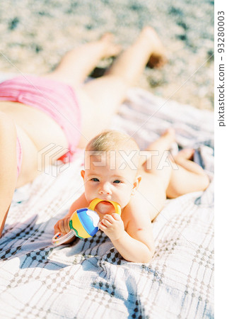 Baby with a rattle in his hands lies next to his mom on a blanket on the beach 93280003