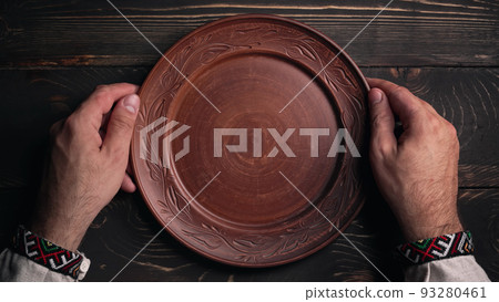 Male hands with empty ukrainian clay plate from wooden table. Man in traditional embroidered shirt 93280461