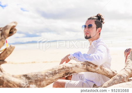 A man wearing Hispanic sunglasses relaxing with his arms on a big tree on a beach in Australia 93281088