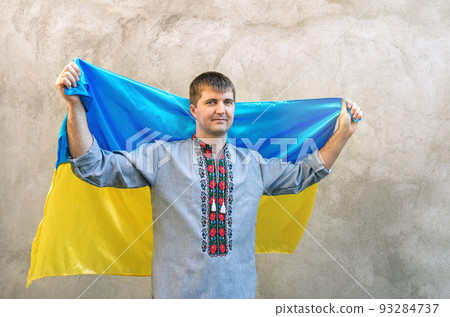 Young man holds blue-yellow flag of Ukraine above his head. Man in traditional embroidered shirt against background of textured concrete wall. Front view, copy space. Independence Day of Ukraine Young man holds blue-yellow flag of Ukraine above his head. Man in traditional embroidered shirt against background of textured concrete wall. Front view, copy space. Independence Day of Ukraine 93284737