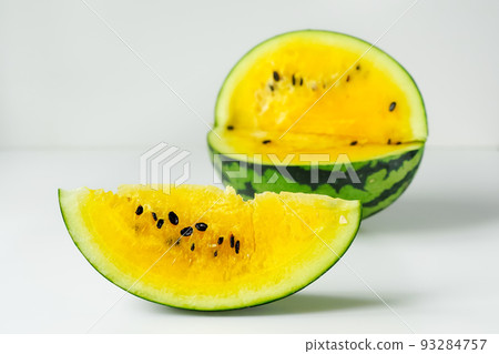 One slice of yellow watermelon in close-up, behind it a blurred oval of a striped giant berry in green rind, on a white background. Isolated slice of watermelon with yellow flesh on light surface. 93284757
