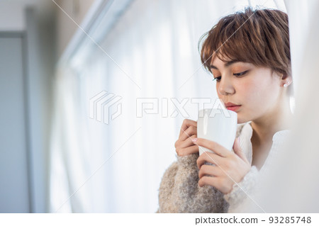 Young woman drinking a hot drink by the window Young woman drinking a hot drink by the window 93285748
