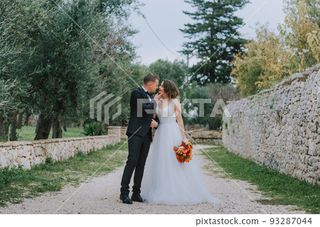 Happy stylish smiling couple walking in Tuscany, Italy on their wedding day. The bride and groom walk down the street by the hands. A stylish young couple walks. Husband and wife communicate nicely Happy stylish smiling couple walking in Tuscany, Italy on their wedding day. The bride and groom walk down the street by the hands. A stylish young couple walks. Husband and wife communicate nicely 93287044