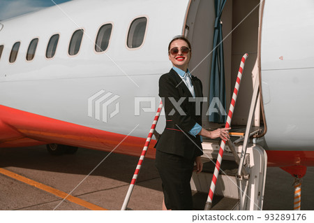 Woman flight attendant in sunglasses standing on airplane stairs at airport and looking away 93289176