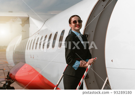 Woman flight attendant in sunglasses standing on airplane stairs at airport and looking camera 93289180