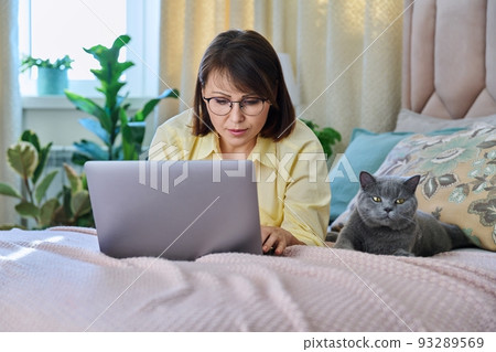 Middle aged woman relaxing at home on bed with laptop and pet cat Middle aged woman relaxing at home on bed with laptop and pet cat 93289569