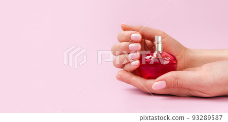 Hands holding a red perfume bottle. Nice female hand with clean and minimalistic manicure with a cosmetic jar. Stylish trendy nails with beauty product on pastel pink background. Front view 93289587