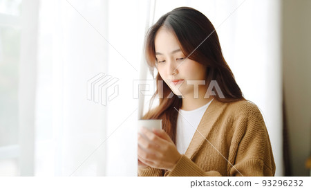 Young asian woman standing beside window and holding mug in living room at home Young asian woman standing beside window and holding mug in living room at home 93296232