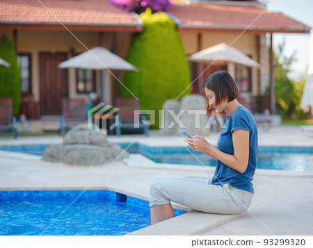 brunette woman using her phone while relaxing by the pool 93299320