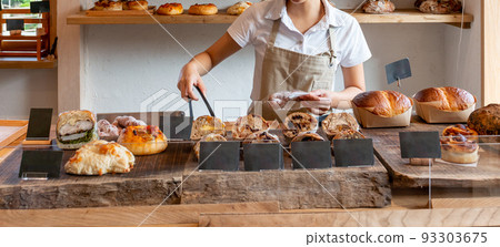 Bakery bakery shop A female clerk in an apron holding freshly baked bread with tongs Bakery bakery shop A female clerk in an apron holding freshly baked bread with tongs 93303675
