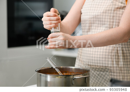 A woman in an apron adds spices to borscht. A woman in an apron adds spices to borscht. 93305948