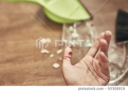 Selective focus on a cut female finger on the background of a split cup and a brush with a dustpan for cleaning. A wound with blood received when cleaning broken dishes. High quality photo 93308919