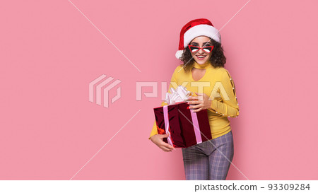 Young smiling happy cheerful curly girl in Santa's hat holds red giftbox while stands on pink background. Sopping Young smiling happy cheerful curly girl in Santa's hat holds red giftbox while stands on pink background. Sopping 93309284