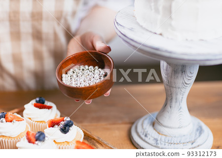 Pastry chef confectioner young caucasian woman in beige apron decorate sweet balls candy cake on kitchen table. Cakes cupcakes and sweet dessert 93312173