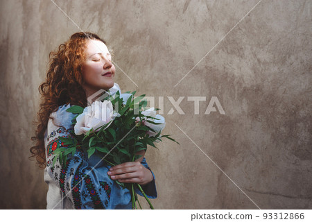 Girl in embroidered shirt bouquet of white peonies. Woman with long red curly hair and closed eyes against concrete wall background. Dark key photo. Independence Day of Ukraine. Copy Space. Soft focus 93312866
