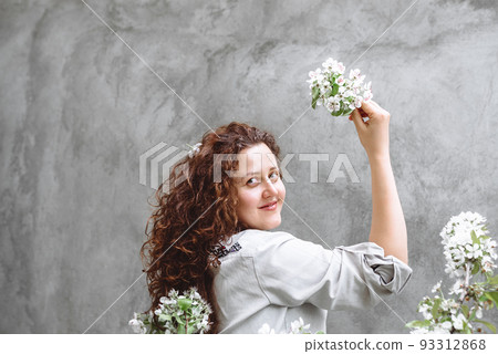 Girl with long curly brown hair in linen shirt holds blossoming branch of apple tree in hand against background of textured concrete wall. Spring flowering time. Copy SpaceIndependence Day of Ukraine Girl with long curly brown hair in linen shirt holds blossoming branch of apple tree in hand against background of textured concrete wall. Spring flowering time. Copy SpaceIndependence Day of Ukraine 93312868