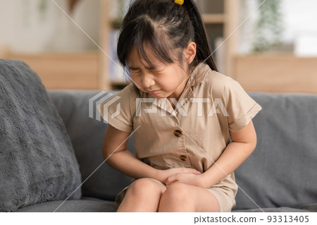 little Asian girl have a stomach ache on couch at home. Child squeeze on her belly so pain and illness. kid has a grimace face. Child health care 93313405
