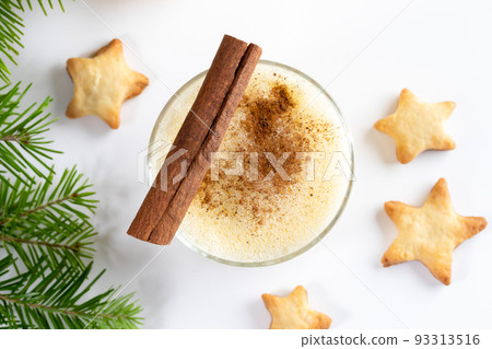 Traditional Christmas treat. Glass of homemade eggnog with cinnamon stick, Xmas cookies stars, fir branches on white background. View from above. Close up. Horizontal orientation. Traditional Christmas treat. Glass of homemade eggnog with cinnamon stick, Xmas cookies stars, fir branches on white background. View from above. Close up. Horizontal orientation. 93313516
