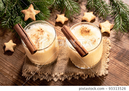 Two glasses of homemade eggnog with cinnamon, fir branches, cookies stars on burlap on rustic wooden table. Traditional Christmas drink. Holiday cozy image. Low key. Back Lit with shadows. Top view. 93313526