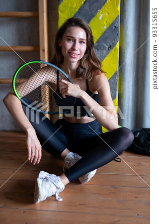 Portrait of female tennis player with racket sitting on gym floor Portrait of female tennis player with racket sitting on gym floor 93315655