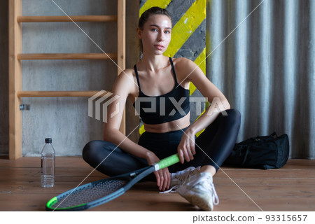 Portrait of female tennis player with racket sitting on gym floor Portrait of female tennis player with racket sitting on gym floor 93315657
